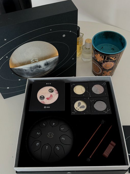An open HEX-themed gift set containing planetary/moon designs, a numbered black disc (1-7), and small containers with celestial-themed items, displayed alongside a candle, perfume bottle, and decorative box.