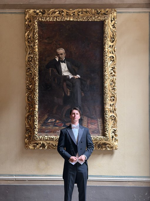 A young man in a black tuxedo stands in front of a large, ornate gold-framed portrait of a seated gentleman in formal attire.