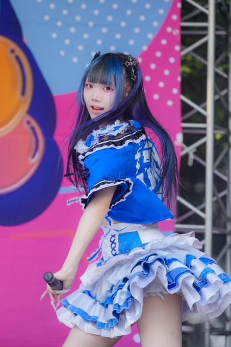 A young female performer with long dark hair and blue-streaked bangs wears an elaborate blue and white frilly idol dress while holding a microphone on stage.