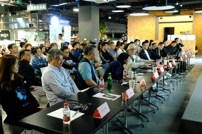 A photograph of an audience seated at a long conference table during what appears to be a panel or industry event, with name placards and water bottles arranged in front of attendees.