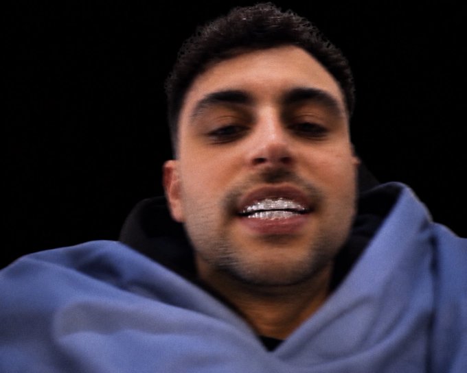 Close-up of a man with short dark hair and a light blue hoodie smiling, showing silver/grill-covered teeth against a black background.