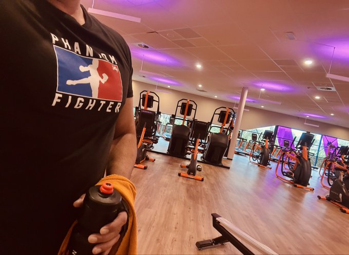 A person in a black 'PHANTOM FIGHTERS' t-shirt holds a water bottle and orange towel inside a Planet Fitness gym featuring purple lighting and orange workout machines.