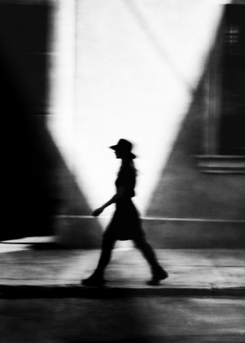 The image features a woman walking down a sidewalk, wearing a hat and dressed in a dress. The photo has been edited with black and white tones, giving it an artistic and dramatic appearance. The woman's silhouette stands out against the background, creating a striking visual effect.
