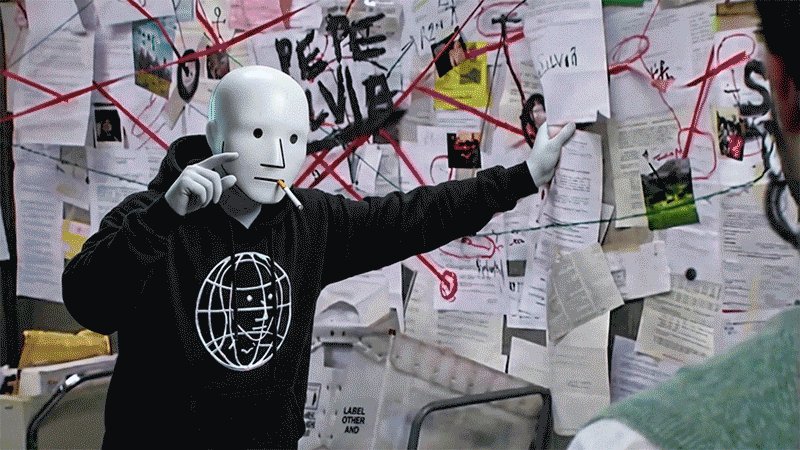 The image shows a person wearing a white mask with a black face, standing against a wall covered in various posters and papers. The person appears to be dressed in a black hoodie or sweatshirt. In front of them, there are several pieces of paper scattered around, possibly related to the content displayed on the posters behind them. The image also contains text that reads "POV: An NPC trying to explain liquid NFTs, mediacoins, memcoins v2.0 and “experiMINTs”". This suggests that there might be a discussion or explanation about digital assets like NFTs (Non-Fungible Tokens) in the context of the image's setting.