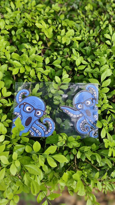 Two blue octopus stickers with glasses peeking out from a clear plastic square, placed on a bed of green leaves.