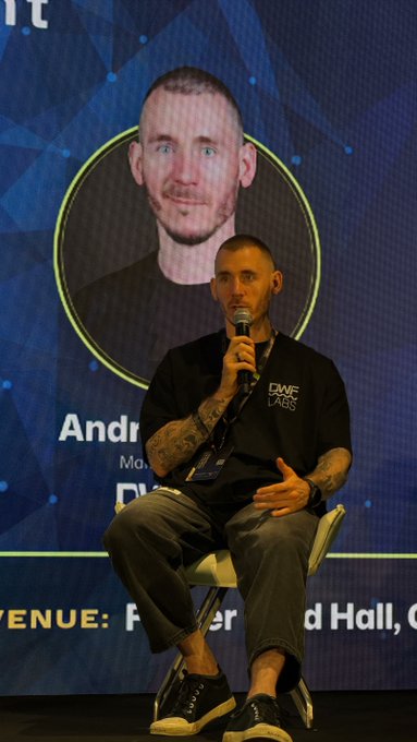 A man with a shaved head, tattoos, and a black t-shirt sits on a chair holding a microphone during a panel discussion, with his image displayed on a screen behind him labeled 'Andre'.