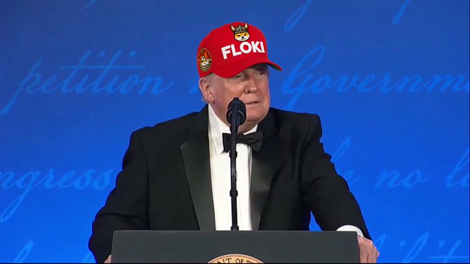 Donald Trump in a tuxedo and bow tie stands at a podium speaking into a microphone, wearing a red 'FLOKI' branded cap.