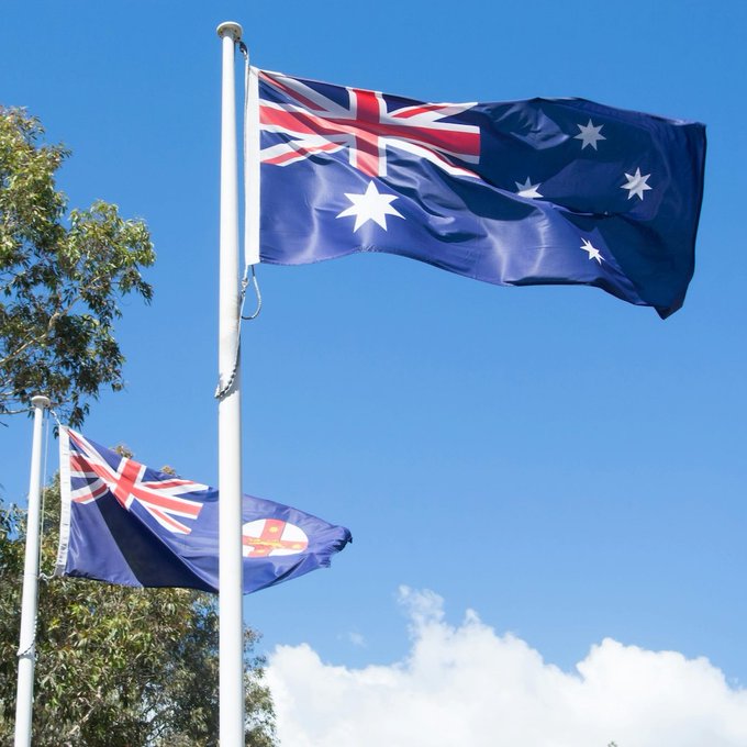 The Australian flag flies prominently against a clear blue sky, symbolizing the nation at the center of ASIC's new regulatory crackdown on digital asset firms. A second flag bearing a state emblem waves below, reinforcing the Australian focus of the September 30 licensing deadline and the threat of fines up to 10% of annual turnover for non-compliance.