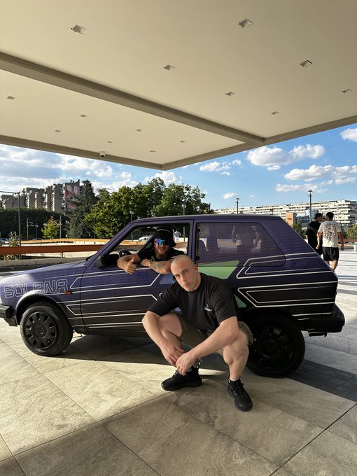 Two men posing with a custom purple Scirocco car featuring horizontal stripe decals, parked under a covered outdoor area with buildings in the background.