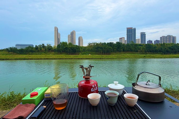 A tea setup with a portable gas canister, kettle, teapot, and small cups arranged on a table overlooking a green river, with a city skyline of high-rise buildings in the background.