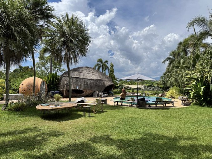 A thatched-roof pavilion and dome-shaped sauna sit poolside amid palm trees, with a round table and lounge chairs arranged on the lawn—offering a tropical "office" where the sauna, ice bath, and coworking setup of Chiang Mai turn a workday into a wellness retreat.