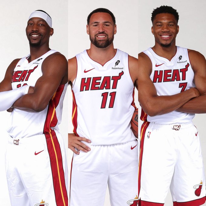 Three Miami Heat players pose in their white "HEAT" jerseys, with the #11 jersey visible on the central player, representing the team's lineup of stars including Bam Adebayo. The image accompanies a quote from Adebayo discussing the Heat's offseason additions of Klay Thompson and Giannis Antetokounmpo as title-contending moves, with the smiling trio visually embodying the "different energy" he describes.