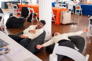 The image shows two men lying down on bean bag chairs with their arms stretched out in front of them. They are positioned near a dining table, which has various items such as books and suitcases scattered around it. The room also contains several chairs placed at different locations throughout the space.