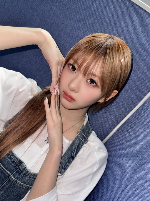 A young woman with long light brown hair and glittery hair accessories poses against a blue wall wearing a white blouse and denim overalls.