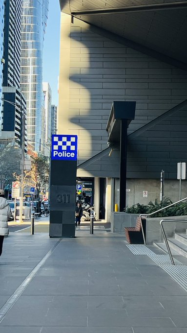 A street-level photo of an urban setting featuring a blue and white checkered 'Police' sign on a stone pillar marked '311', with tall buildings, a pedestrian in a white jacket, and street elements visible in the background.
