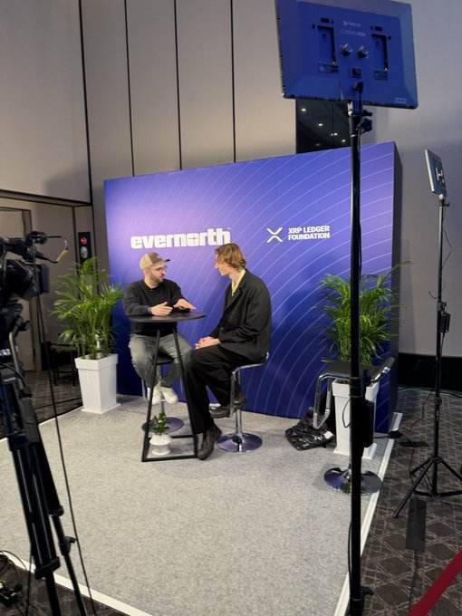 A behind-the-scenes look at an interview setup featuring two men seated at a table in front of an "evernorth" and "XRP LEDGER FOUNDATION" branded backdrop, with professional lighting and camera equipment visible around the set. The scene connects to the caption "The real @Vet_X0" by showing what appears to be an XRP Ledger Foundation interview with the individual known by that handle.