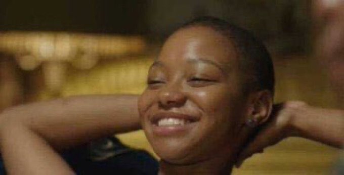 A young woman with short hair is laughing joyfully, her head tilted back and hands clasped behind her neck in a relaxed pose.