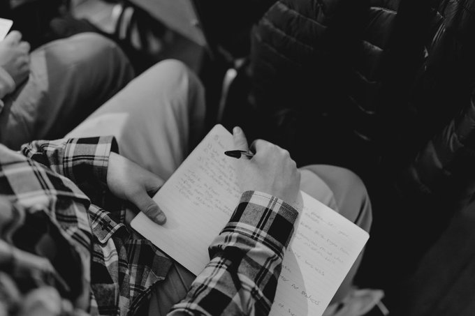 A person in a plaid shirt writes notes in a notebook on their lap, embodying the message that unseen dedication—like quiet study and reflection—eventually leads to visible rewards. The handwritten page reflects the private effort behind future accomplishments.