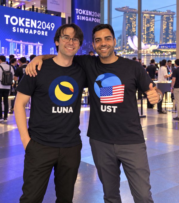 Two men posing together at the TOKEN2049 SINGAPORE event, both wearing black t-shirts—one with a 'LUNA' logo and the other with 'UST' and an American flag design—with the Singapore skyline visible in the background.