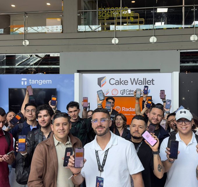 A group of people pose together holding up smartphones, standing in front of banners for 'Cake Wallet', 'Cake Labs', and 'tangem' at what appears to be a tech or crypto event.