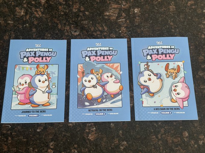 Three comic books titled 'Adventures of Pax Pengu & Polly' rest on a dark surface, featuring Volume 1 'Journey to the Berg', Volume 2 'Betrayal on the Berg', and Volume 3 'A New Dawn on the Berg'.