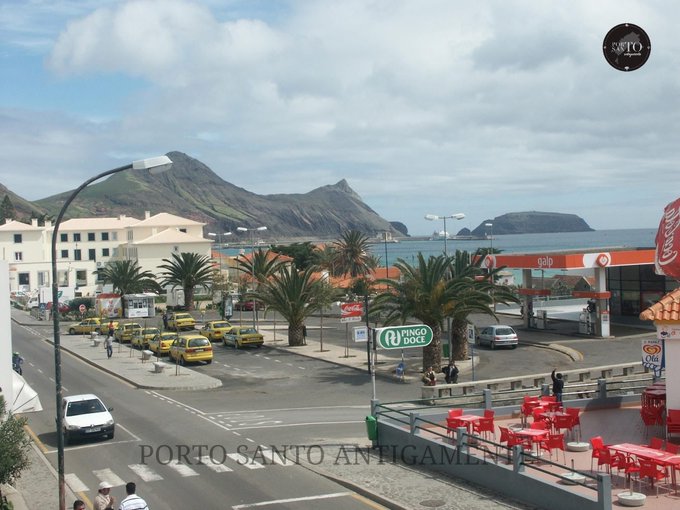 A scenic coastal view of Porto Santo Antigo features yellow taxis parked along a street lined with palm trees, a Galp gas station, and mountains rising from the sea in the background.
