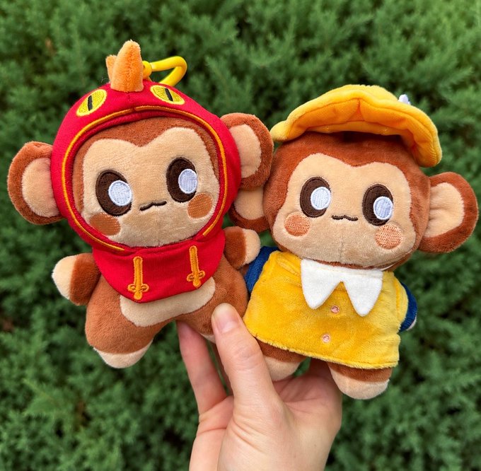 A hand holding two plush monkey toys outdoors against green foliage—one wearing a red dragon-style costume and the other in a yellow hat and bib outfit.