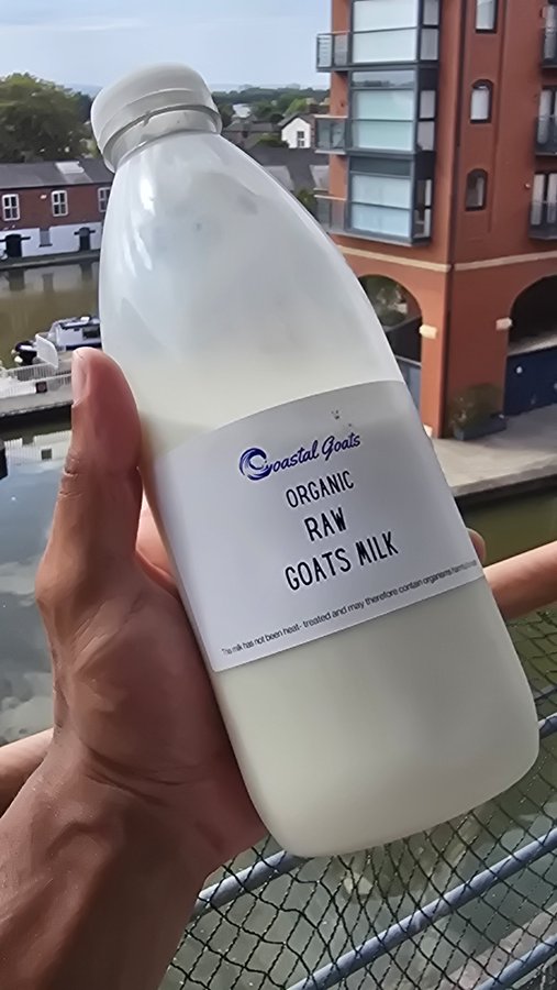 A hand holds up a glass bottle of "Coastal Goals" Organic Raw Goats Milk, with a view of brick apartment buildings and a waterway in the background.