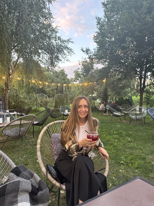 A young woman with long blonde hair smiles while sitting in a wire chair at an outdoor venue, holding a glass of red drink, with string lights and greenery in the background.