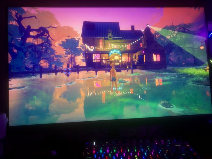 The image shows a computer monitor displaying a video game scene with a house and people standing by a lake or pond. The screen has a purple hue that adds an artistic touch to the overall presentation. In addition, there are two keyboards visible on the desk in front of the monitor, suggesting that this setup is likely used for gaming purposes.