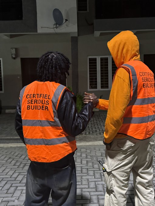 Two people wearing orange high-visibility vests; one vest reads 'CERTIFIED SERDUS BUILDER' and the other reads 'CERTIFIED SERIOUS BUILDER', standing outside at night near a building with a satellite dish.