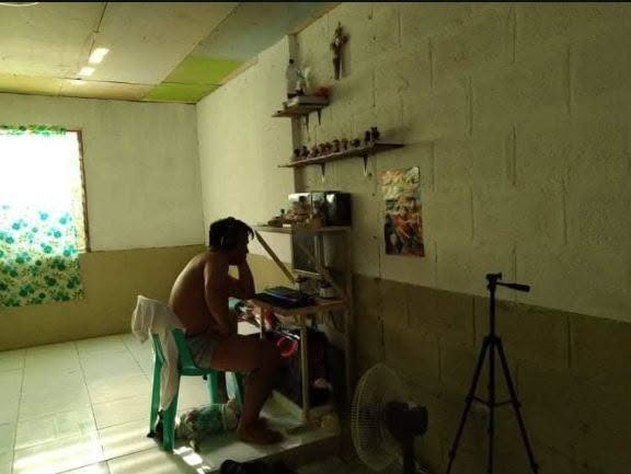 A shirtless man sitting on a green plastic stool in a room with concrete block walls, appearing to work on a piece of electronic equipment on a desk, with a tripod and fan nearby.