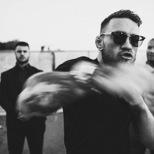 A black and white photo of a bearded man wearing sunglasses and a black shirt, gesturing with his hands blurred in motion, with two other men standing in the background.