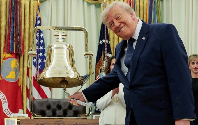 Donald Trump smiles broadly while pulling the rope to ring the Liberty Bell in the Oval Office, with American and Marine Corps flags visible in the background.