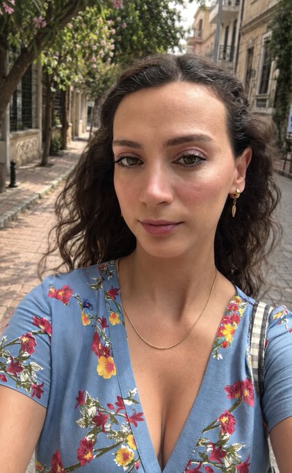 A selfie of a woman with dark curly hair and pink eyeshadow, wearing a blue floral dress and a delicate gold necklace, standing on a sunny street.