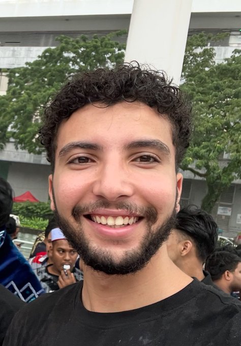 A smiling young man with curly hair poses for a selfie outside a building, with a crowd visible behind him. The caption suggests this is him in Tunisia, sharing a hopeful self-introduction after losing his crypto savings in early 2025 and choosing to start learning again.