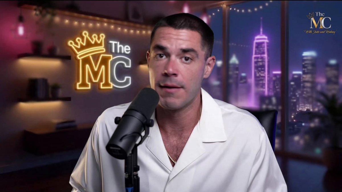 !"The MCC" is written above a microphone, which is held by a man wearing a white shirt. The background of the image shows a city skyline at night with buildings lit up against the dark sky.