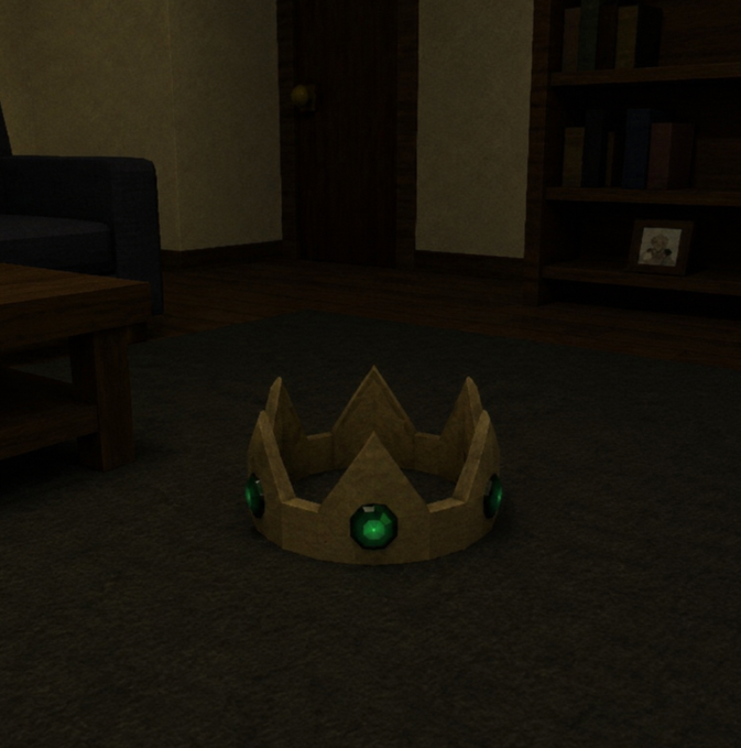 A cardboard crown with green gemstones rests on a dark carpet in a dimly lit room with a coffee table and bookshelf in the background.