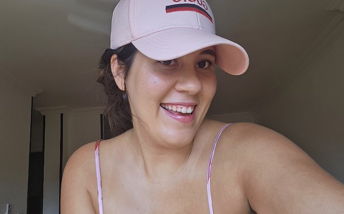 A smiling woman wearing a pink baseball cap and a pink spaghetti strap top takes a selfie indoors.