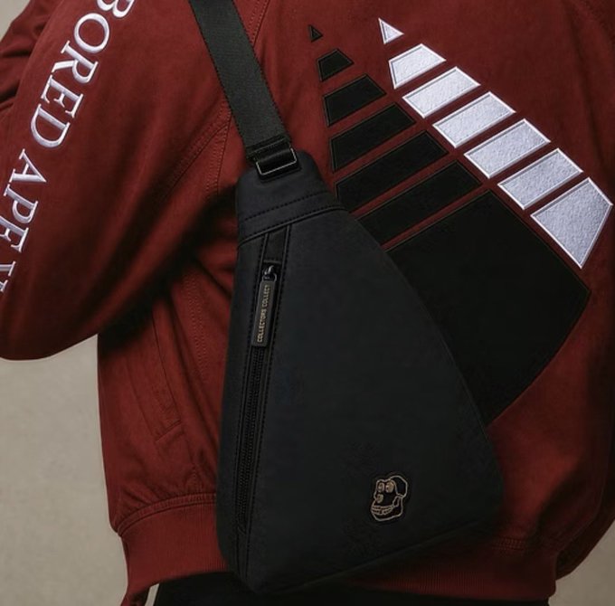 A close-up photo of a person wearing a dark red jacket with 'BORED APE' text and graphic patches, carrying a black triangular sling bag with a small gorilla/monkey logo and zipper detail.