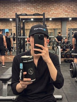 A man in a black cap and shirt takes a mirror selfie in a gym while holding up a black ribbed tumbler branded with "Trade smarter" and a "Bitget" sticker, showcasing the merchandise gift. The post expresses gratitude to @KT_BTC for the received item, matching the Bitget-themed cup shown.
