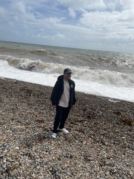 A person in a black jacket and cap stands on a pebbled beach as choppy, white-capped waves roll in from the sea behind them.
