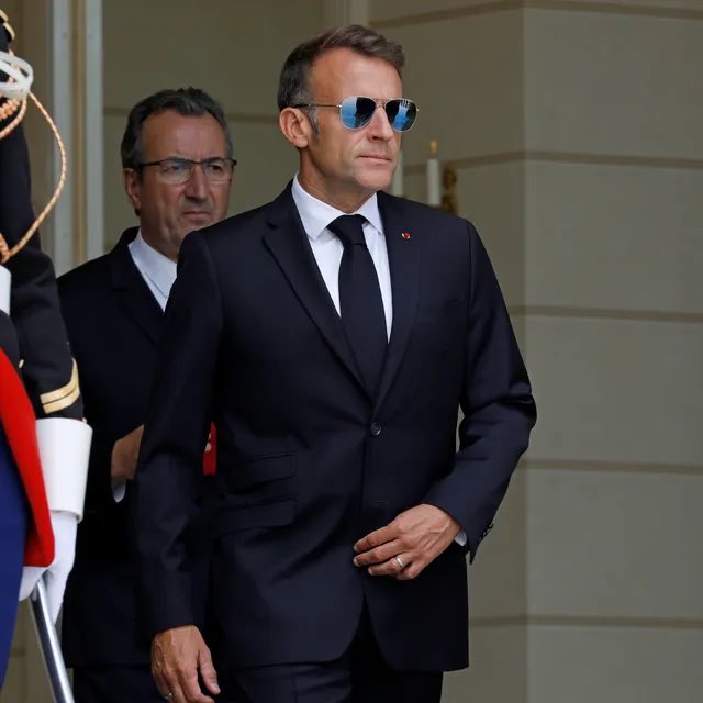 Photograph of French President Emmanuel Macron wearing aviator sunglasses and a dark suit, walking alongside another man in a suit at what appears to be an official ceremony.