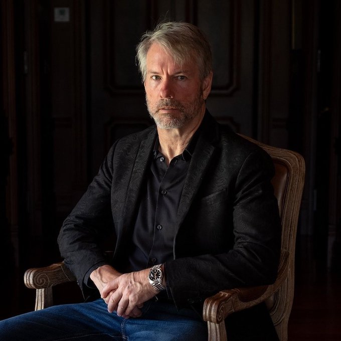A distinguished man with graying hair and a beard, wearing a black patterned blazer over a dark shirt and jeans, sits in a wooden chair looking directly at the camera. The accompanying caption connects the image to Michael Saylor, a prominent Bitcoin advocate known for his views on converting economic energy into digital form.