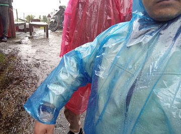 Two people wearing clear plastic rain ponchos, one blue and one red, stand outdoors on a wet, muddy path. The blue poncho has 'SMC' printed on the sleeve.