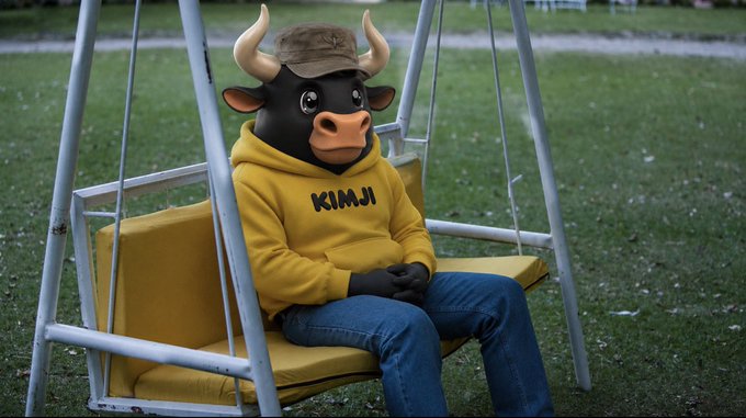 A person in a black bull mascot costume wearing a yellow hoodie emblazoned with "KIMJI" sits pensively on a yellow swing in a grassy field, embodying the post's message of patient bullish optimism as "the wait will soon be over."