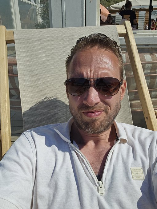 A man wearing sunglasses relaxes in a wooden beach chair, embodying the carefree beach lifestyle described in the post while his investments in $OMHD and $APEX climb in value. The sun-drenched scene perfectly captures the sentiment of earning money effortlessly while enjoying leisure time.
