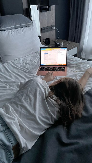 A cozy lifestyle photo of a person with long hair lying in bed, partially covered by white sheets, using a laptop displaying what appears to be a spreadsheet.