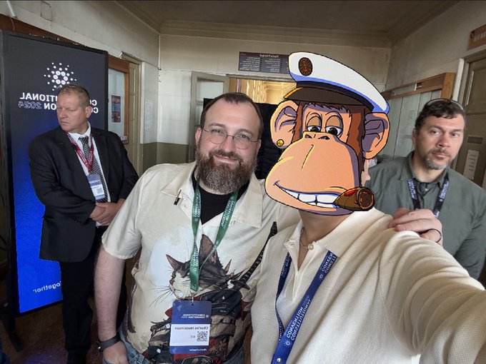A group selfie taken at what appears to be a conference, featuring a smiling bearded man in a white shirt with lanyards, another man in a suit on the left, and a person on the right whose face is obscured by a cartoon monkey captain emoji.