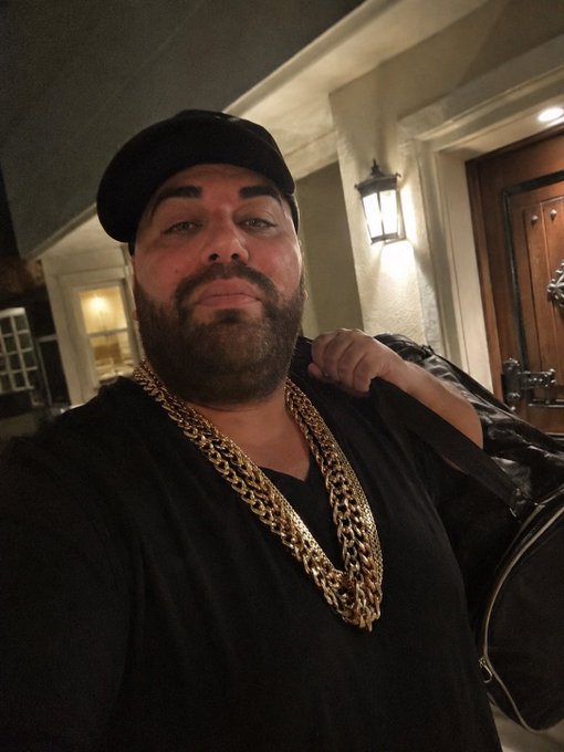 A bearded man wearing a black beanie and multiple gold chains poses for a selfie outside a home's front door, dressed in dark clothing while carrying a bag over his shoulder. The image humorously personifies the suspicious-looking "Trezor Customer Support" character mentioned in the caption, playing on the joke that someone from a crypto hardware wallet company showing up unannounced to verify your delivery would look more like a gangster than a legitimate tech representative.